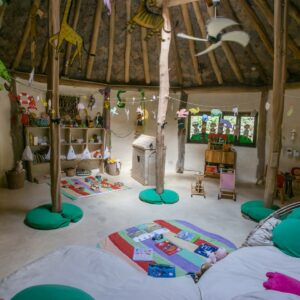 A cozy, rustic children&#039;s playroom with wooden beams, a round thatched roof, and various toys and books neatly arranged. Colorful decorations hang from the ceiling, with cushions and play mats on the floor. In the center stands a small playhouse, embodying the charm of Soneva Exclusive Offers.