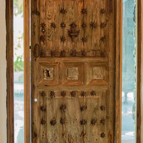 A rustic wooden door adorned with metal star-shaped decorations and carved panels stands open, framed by glass panels on each side. The door shows signs of age and weathering, adding character to its appearance. Sunlight filters through the glass, illuminating the doorway, reminiscent of a Maldives exclusive experience at Soneva.