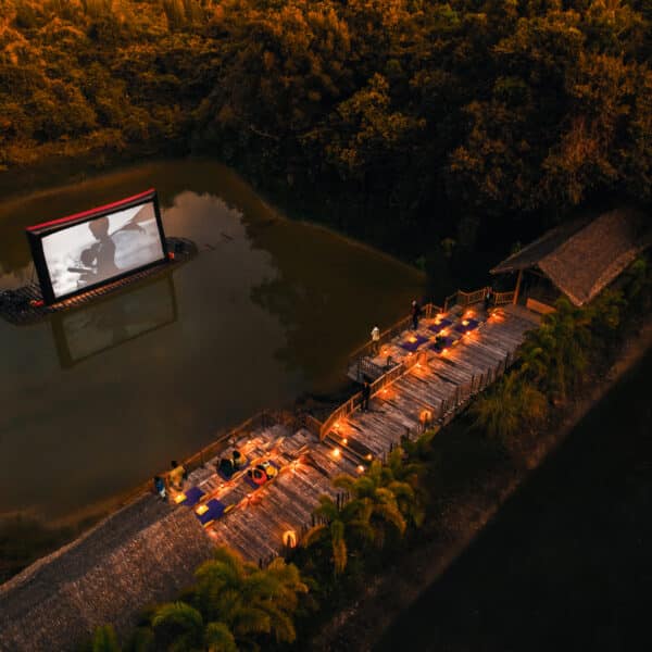 Aerial view of an outdoor movie setting at dusk. A large screen displays a black-and-white film over a small lake, with seating on a wooden pier lined with lights leading from the shore. Surrounded by dense trees, this exclusive experience at Soneva in Thailand creates a serene and inviting atmosphere.