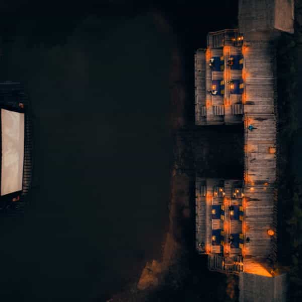 Aerial view of a floating outdoor cinema at night in Thailand. The screen is mounted on a boat in a body of water, facing a seating area on the shore made of wooden platforms. The platforms have chairs and are softly lit by numerous small lights, creating a serene and inviting ambiance for this exclusive Soneva experience.