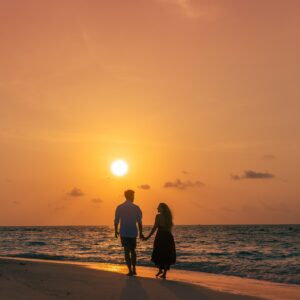 A couple holds hands while walking along a beach at sunset, their forms silhouetted against the orange and pink hues of the sky. The ocean waves gently lap at the shore to their right, creating a scene that could easily be part of Soneva Exclusive Offers.