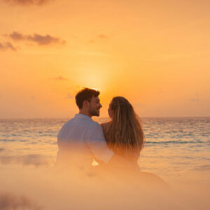 A couple sits on the beach, embracing and looking out at the ocean during a vivid orange sunset. The sky is filled with soft, scattered clouds, and the serene waves add to the romantic and peaceful atmosphere, reminiscent of Soneva Exclusive Offers&#039; enchanting getaways.