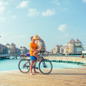 A couple embraces next to a bicycle on a wooden pier over turquoise water. Wooden bungalows on stilts are visible in the background under a sunny, blue sky. One partner wears an orange shirt and blue shorts, and the other wears a floral shirt and sunhat, enjoying their Soneva Exclusive Offers vacation.