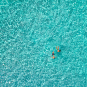 Aerial view of two people swimming in clear, turquoise water at a Soneva resort. The sunlight reflects off the water, creating a pattern of ripples and waves. The swimmers are side by side, enjoying the pristine, refreshing environment amidst exclusive offers from Soneva.