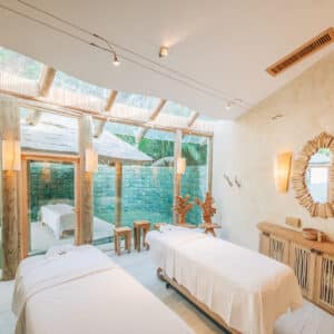 A serene Maldives spa room with two massage tables covered in white sheets. The room features natural wooden furniture, soft lighting, and a large window offering a view of an outdoor area with greenery and a small pool. Walls are painted in a light, calming color, enhancing the wellness experience.