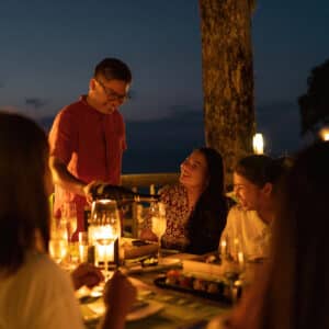 A group of people are enjoying an outdoor dinner by candlelight at dusk during a Thailand exclusive experience. A man in a red shirt is standing and pouring a drink while others, seated around the table, are smiling and engaged in conversation. The ambiance is warm and serene with lights and a tree in the background, echoing the luxurious essence of Soneva.