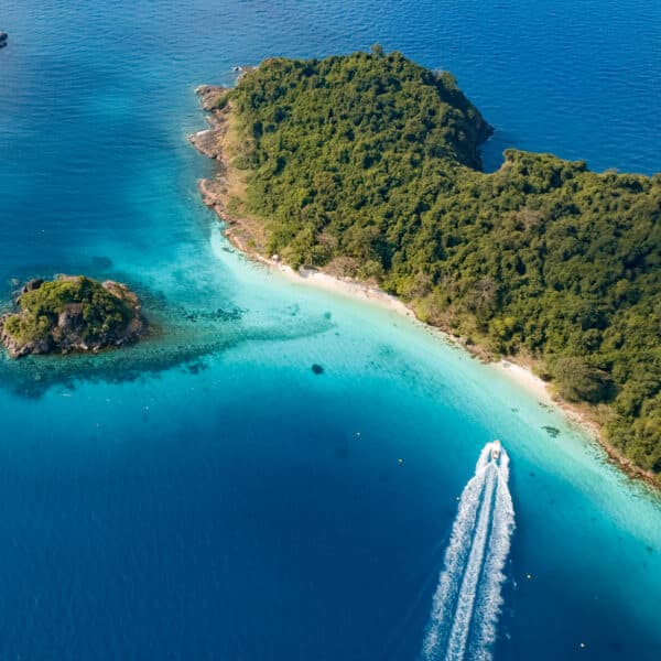 Aerial view of a tropical island with lush greenery surrounded by clear, turquoise waters. A speedboat with a trail of white wake is moving near the shoreline. Two smaller, green-covered islets are visible nearby. The overall scene is vibrant and serene—an exclusive experience reminiscent of Soneva Thailand.