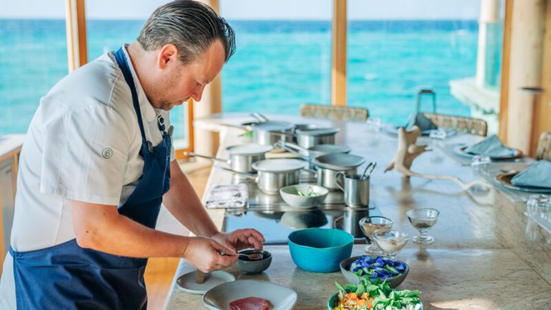 A chef in a white uniform and blue apron prepares a dish at a kitchen counter with various ingredients and pans. The kitchen, part of the luxurious Maldives dining experience at Soneva Fushi, has large windows offering a view of the ocean, creating a bright and airy atmosphere.