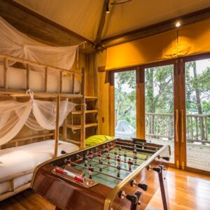 A cozy treehouse interior features wooden bunk beds with mosquito nets, a foosball table, and bean bags. Natural light floods in through large windows, offering a scenic view of the surrounding forest and a wooden balcony. Take advantage of Soneva Exclusive Offers for a unique retreat.