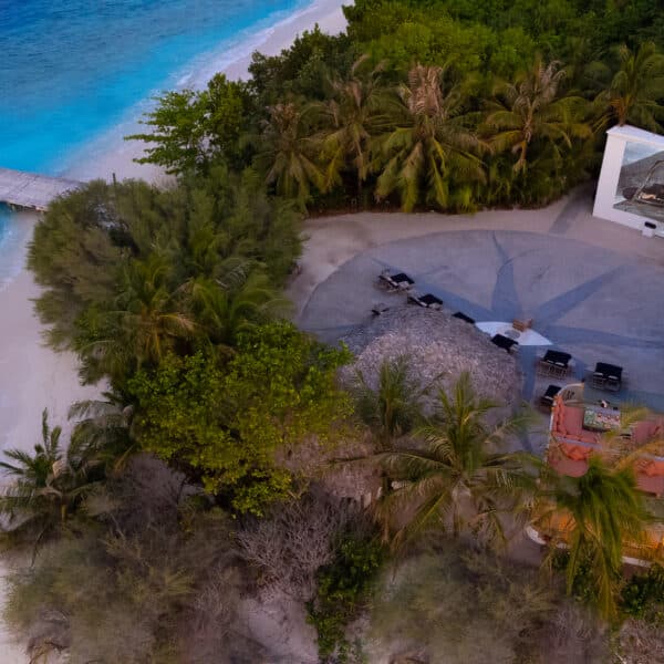 Aerial view of a beachside outdoor cinema at Soneva, offering an exclusive Maldives experience. A large screen displays a movie, surrounded by trees and facing crystal-clear blue waters. A wooden walkway extends over the water to the shore, where several lounge chairs are arranged for viewers.