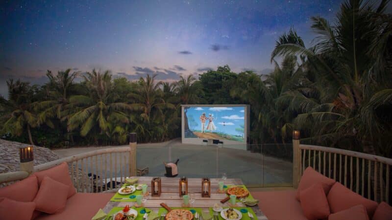 A cozy outdoor dining setup with a green table and cushioned seating, featuring a variety of dishes and lanterns. Behind the dining area, a large screen displays an animated movie. The background has a starry night sky and surrounding palm trees, creating a serene Maldives exclusive experience.