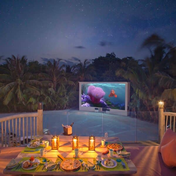 A cozy outdoor dining setup with a table adorned with candles, plates of food, and drinks. The table overlooks a lush, tropical landscape under a starry night sky. A television screen in the background is playing an animated movie featuring a clownfish—truly a Maldives Exclusive Experience at Soneva.