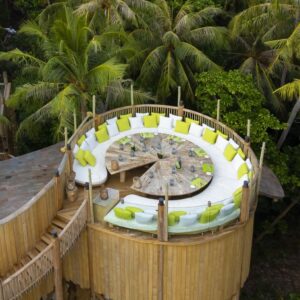 Aerial view of a circular wooden treehouse nestled among palm trees. The treehouse, part of Soneva Exclusive Offers, has an open rooftop lounge area with cushioned seating in lime green and white surrounding a central wooden table. A staircase leads up to the treehouse through lush greenery.