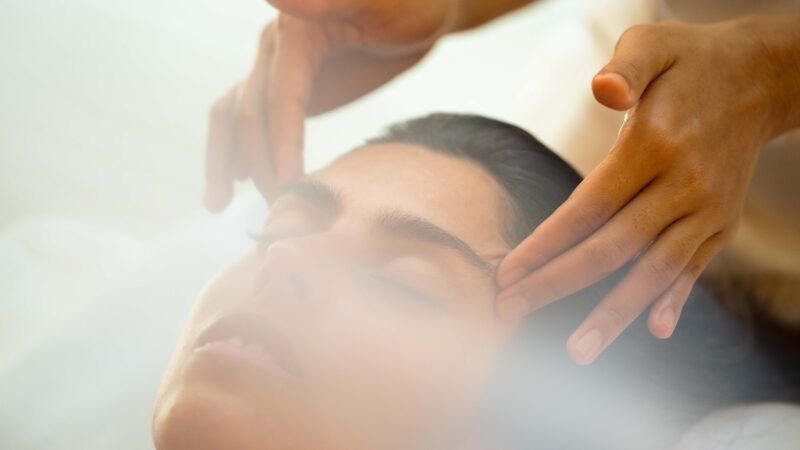 A person with eyes closed receives a gentle facial massage at a Maldives spa. Two hands are visible applying light pressure to the temples and forehead. The background is soft and blurry, emphasizing the relaxed wellness ambiance.