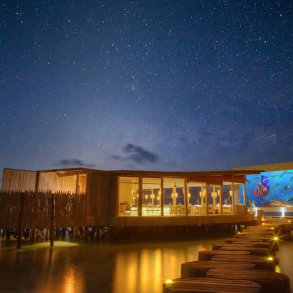 A stunning overwater restaurant at Soneva, illuminated at night under a starry sky. A long wooden walkway leads to the exclusive Maldives experience, with calm water reflecting the lights. A colorful mural depicting a marine scene adorns one side of the structure.