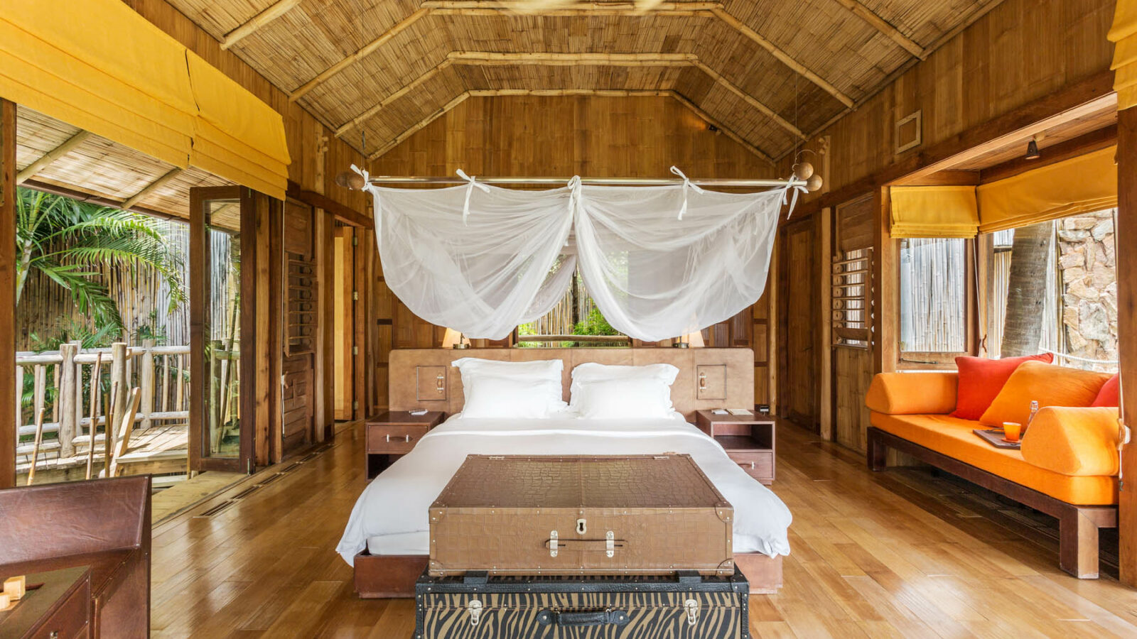 A spacious wooden bedroom with a high ceiling, featuring a white-draped canopy bed, orange sofa, and wooden furniture. Large windows and doors let in natural light, offering views of the lush greenery outside. Perfect for a Thailand family holiday at Soneva, a ceiling fan spins overhead.