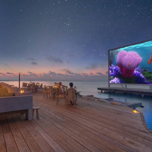 A serene oceanfront setting at night in the Maldives. People sit comfortably on a wooden deck with cozy seating, watching a movie on a large screen. The sky is starry, and the water is calm. This exclusive Soneva experience features an animated under-the-sea adventure, making it unforgettable.