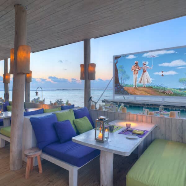 A cozy, beachside restaurant features cushioned booth seating with blue and green pillows along a wooden deck. An outdoor screen displays a vibrant image of a couple in white attire and a bird above them. The background shows an ocean view during sunset, capturing the essence of a Maldives Exclusive Experience.