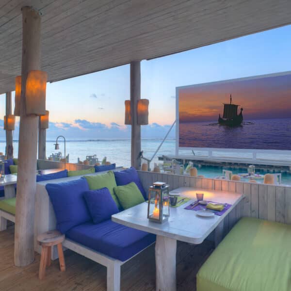 A seaside restaurant with wooden furniture and vibrant green and blue cushions. Tables are set with candles and place settings. The view includes a sea with a large screen displaying a sunset scene of a boat on the water. Lanterns hang from wooden pillars, offering an exclusive Maldives experience reminiscent of Soneva's luxury.