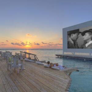 A stunning seaside setup features an outdoor cinema screen showing a black-and-white movie, placed on a pier extending over calm waters. Wooden lounge chairs and tables line the deck, with a vibrant sunset and scattered clouds in the background—a perfect scene showcasing Soneva Exclusive Offers.