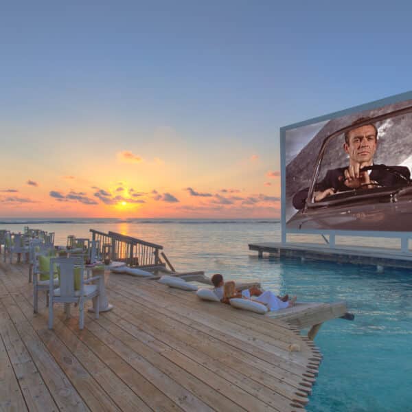 A serene sunset scene over the ocean with a large outdoor movie screen on the water showing a man in a car. Wooden decking with seating and lounge areas extends over the water, with a couple relaxing on cushions, enjoying the view and the movie—truly a Maldives Exclusive Experience at Soneva.