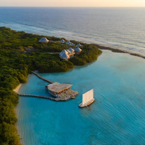 Aerial view of a tropical coastal resort featuring lush greenery and a tranquil turquoise sea. A wooden pier extends into the water with a sailboat docked nearby. Several huts with thatched roofs are nestled among the foliage, offering an exclusive Maldives experience with Soneva at sunset on the horizon.