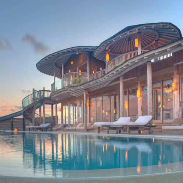 A luxurious waterfront villa at sunset features a large, curved infinity pool with sparkling clear water. The modern wooden structure has large glass doors and windows, sleek outdoor lounge chairs, and a spiral slide leading into the pool from the upper balcony—discover all this with Soneva Exclusive Offers.