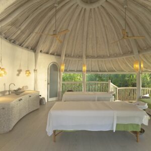 A tranquil spa room with a domed wooden ceiling and large windows revealing a lush green outdoor view. The room features two massage tables with white linens, hanging pendant lights, a sink, and a wooden door. Experience the serene and natural ambiance with our Soneva Exclusive Offers.