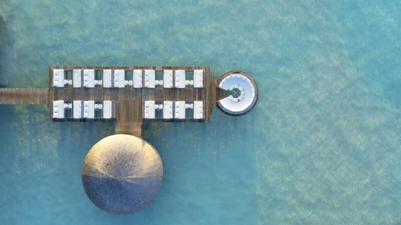 Aerial view of a luxurious overwater bungalow with sunbeds arranged along a wooden deck, leading to a circular open space with a seating area. The entire structure is located in clear, shallow turquoise water, creating a serene and tropical ambiance—perfect for an unforgettable Soneva Maldives holiday.