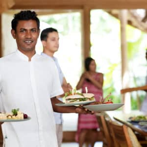 A waiter in a white uniform is holding two plates of food, featuring a variety of dishes, and smiling. In the background, a few other people, including another waiter and some diners, are visible in a well-lit restaurant setting with wooden decor—a day in the life of a Soneva Career.