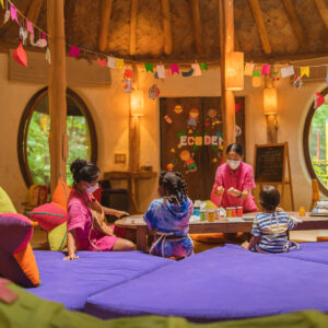 A cozy, circular room with wooden beams and colorful decor feels like celebrating Chinese New Year at Soneva in the Maldives. Four children sit on purple cushions, engaged in arts and crafts. Two adults assist them, while hanging decorations and vibrant, round windows add to the warm, playful atmosphere.