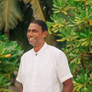 A man stands outdoors, smiling and wearing a white collared short-sleeve shirt. The background is lush with green leaves and palm fronds, suggesting a tropical environment—the perfect setting for someone pursuing a Soneva career.