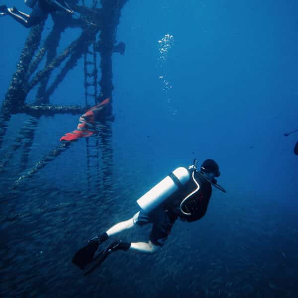 A diver in full gear swims underwater near a large metal structure that extends upwards to the surface. Small fish surround the diver, creating a dense aquatic environment. Nearby, another diver in a red wetsuit and fins explores this Thailand Exclusive Experience, hosted by Soneva.