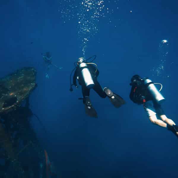 Two scuba divers swim underwater near a sunken shipwreck off the coast of Thailand, with one diver in the background. The divers, enjoying this exclusive experience from Soneva, are equipped with oxygen tanks and flippers as bubbles rise to the surface in the blue ocean water surrounding them.