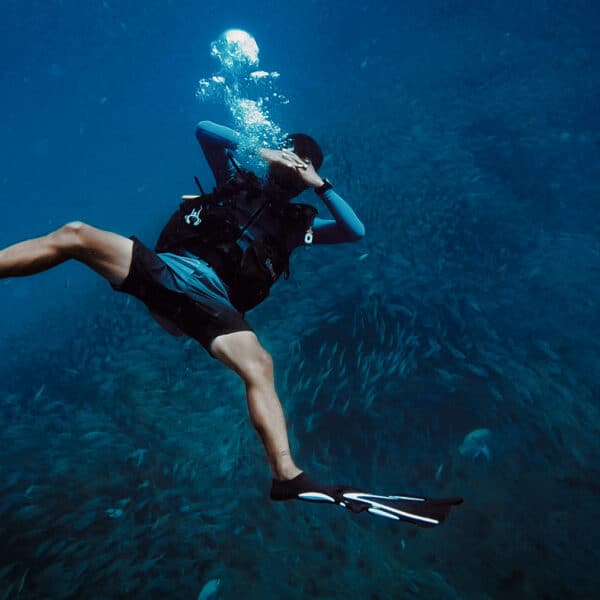 A scuba diver swims underwater surrounded by a school of fish in this exclusive experience at Soneva, Thailand. The diver is wearing fins, a scuba mask, and a wetsuit. Bubbles rise above the diver, and the deep blue ocean creates a serene backdrop.