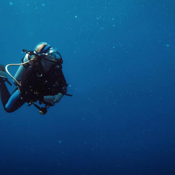 A scuba diver wearing full gear is submerged in deep blue water, enveloped by bubbles and particles. Exploring the underwater environment with fins, oxygen tank, and mask clearly visible, they enjoy a Thailand exclusive experience. The background is a vast, serene expanse of the ocean.