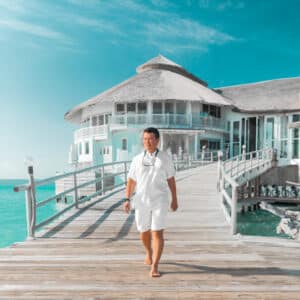 A person in a white outfit walks barefoot along a wooden pier leading to a circular, multi-story beach house with large windows and a thatched roof. The weather is clear and sunny, and the turquoise sea surrounds the scene, reflecting the idyllic lifestyle promoted by Soneva Career.