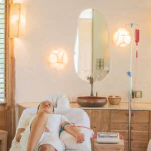 A person receiving an intravenous (IV) therapy treatment while lying on a comfortable bed in a wellness clinic reminiscent of a Maldives spa. The room has soft lighting, wooden furniture, and decorative wall lights. The person is relaxed with their eyes closed and appears to be at ease.