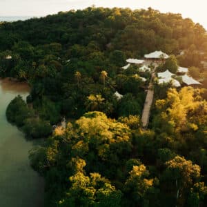 Aerial view of a lush, green tropical island at sunset. Dense jungle vegetation covers the hill, leading down to a calm, clear body of water. Light-colored structures, possibly tents or pavilions from the Soneva Kiri Resort, are nestled among the trees, connected by a walkway.