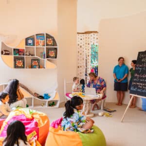 A group of children sit on colorful bean bags in a brightly lit room with a whale-shaped bookshelf. A woman reads from a book while another adult stands near a blackboard with writing. The room has a playful and cozy atmosphere, reminiscent of the charm you'd find with Soneva Exclusive Offers, featuring curtains and natural light.