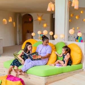 A woman and two young girls sit on an indoor sofa with yellow and green cushions. The woman reads a book to the older girl while the younger one, sitting on her other side, points at something exciting. The room is adorned with soft lighting and playful elements, creating a scene reminiscent of Soneva Exclusive Offers.