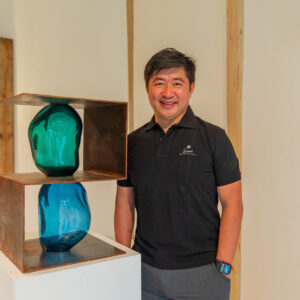 A man in a black polo shirt stands next to a modern art sculpture featuring two blue glass shapes enclosed in a rectangular metal frame. The background, reminiscent of a Maldives exclusive experience at Soneva, includes wooden panels and a wall-mounted light fixture. The man is smiling with his hands by his sides.