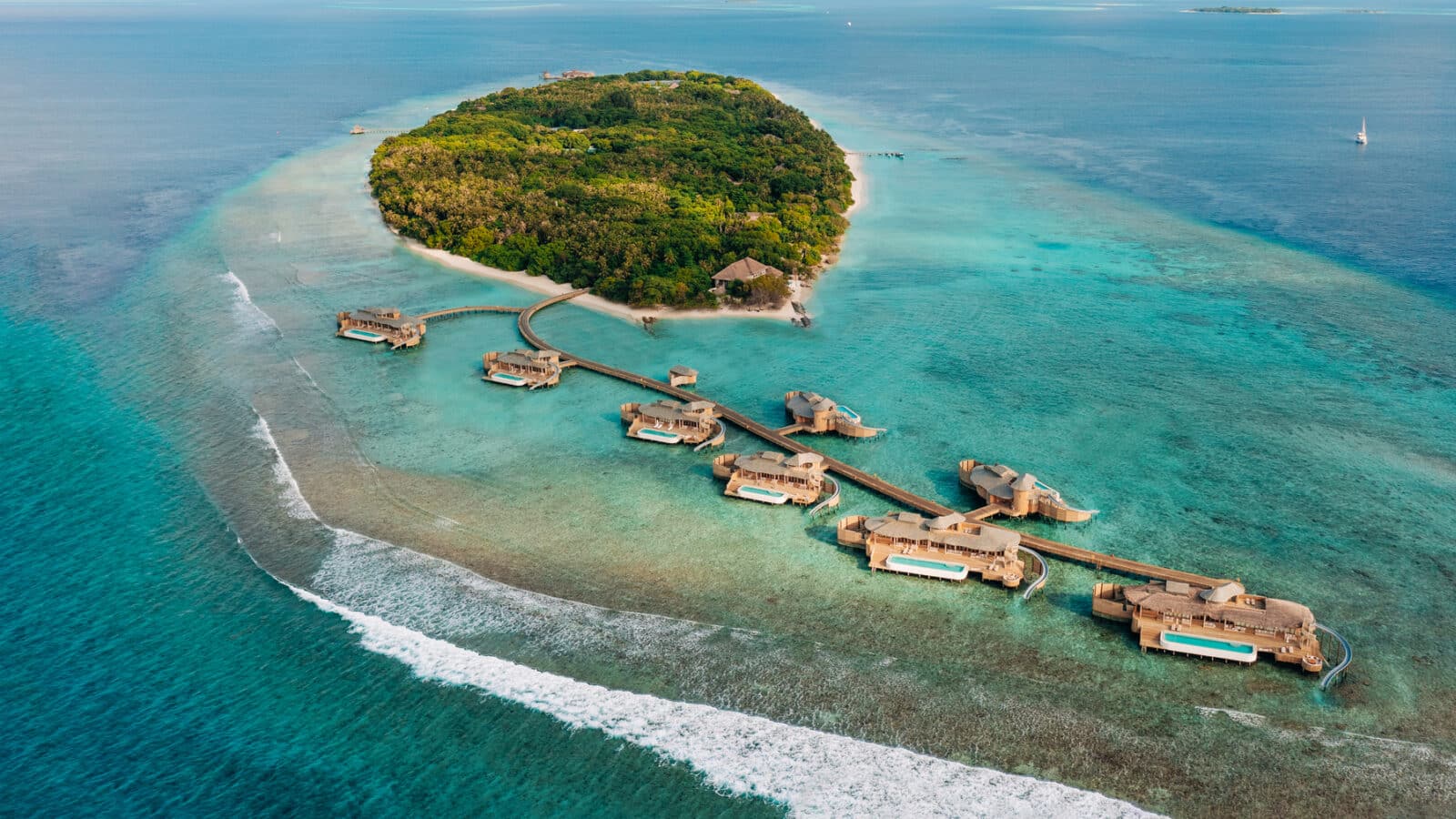 Aerial view of a tropical island surrounded by clear turquoise waters with several overwater bungalows connected by a wooden walkway. The island is lush with greenery, and white sand beaches line the shore. Waves crash gently onto a nearby reef, showcasing Soneva Exclusive Offers' paradise.