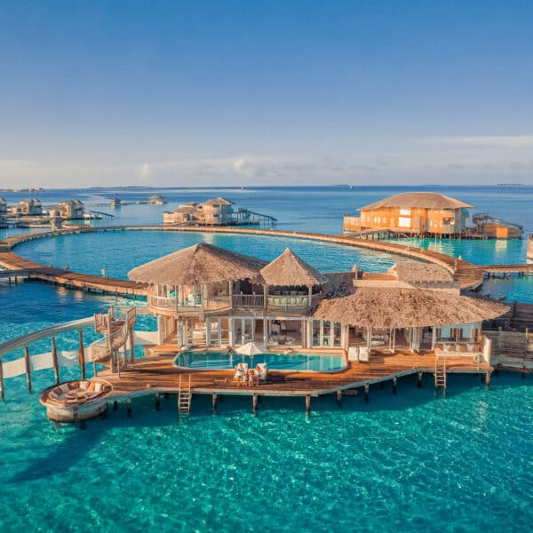 Aerial view of luxurious overwater bungalows with thatched roofs in a Maldives luxury resort. The crystal-clear turquoise sea surrounds the wooden structures connected by boardwalks, some featuring slides that lead directly into the water. Clear blue sky in the background adds to the picturesque Soneva Villas scene.