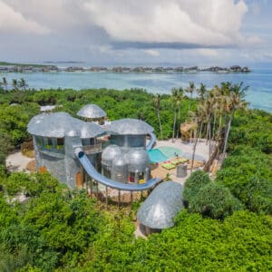 Aerial view of a luxurious treehouse villa with silver domed roofs surrounded by lush green foliage. The villa features an outdoor pool and a slide leading to the pool. In the background, overwater bungalows are visible over turquoise ocean waters under a partly cloudy sky—an example of Soneva Exclusive Offers.