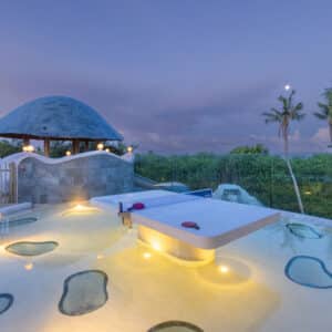 A serene outdoor pool area at twilight, featuring a shallow pool with submerged lounge tables and benches illuminated by soft lights. A thatched-roof gazebo is situated nearby, surrounded by lush greenery and tall palm trees under a dusky sky—a perfect setting for a family holiday at Soneva in the Maldives.