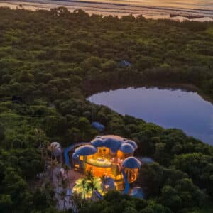 Aerial view of a unique, dome-shaped building nestled within dense greenery near a serene body of water. The structure glows warmly from within, contrasting with the dark foliage, as you envision your perfect family holiday. A calm ocean is visible in the background under a golden sunset sky.