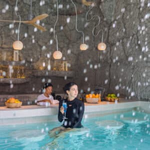 A person stands in a pool grotto at Soneva in the Maldives, holding a camera while water falls around them. In the background, another person is seated at a bar counter with fruits and candles on it. Pendant lights hang overhead, and the walls have a rocky texture, perfect for a serene family holiday.