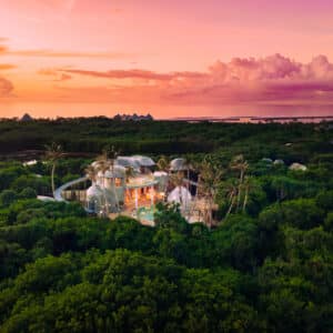 Aerial view of Soneva, a tropical beach resort in the Maldives, perfect for a family holiday, surrounded by lush greenery at sunset. The resort features numerous thatched-roof buildings and structures interconnected by pathways. The sky is a vibrant mix of pink, orange, and purple hues with scattered clouds.