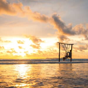 A person swings on a wooden swing suspended over the shore, under a vibrant golden sunset. The sky is dotted with clouds, and the ocean waves gently lap at the beach, reflecting the warm hues of the setting sun—a perfect scene reminiscent of Soneva Exclusive Offers.